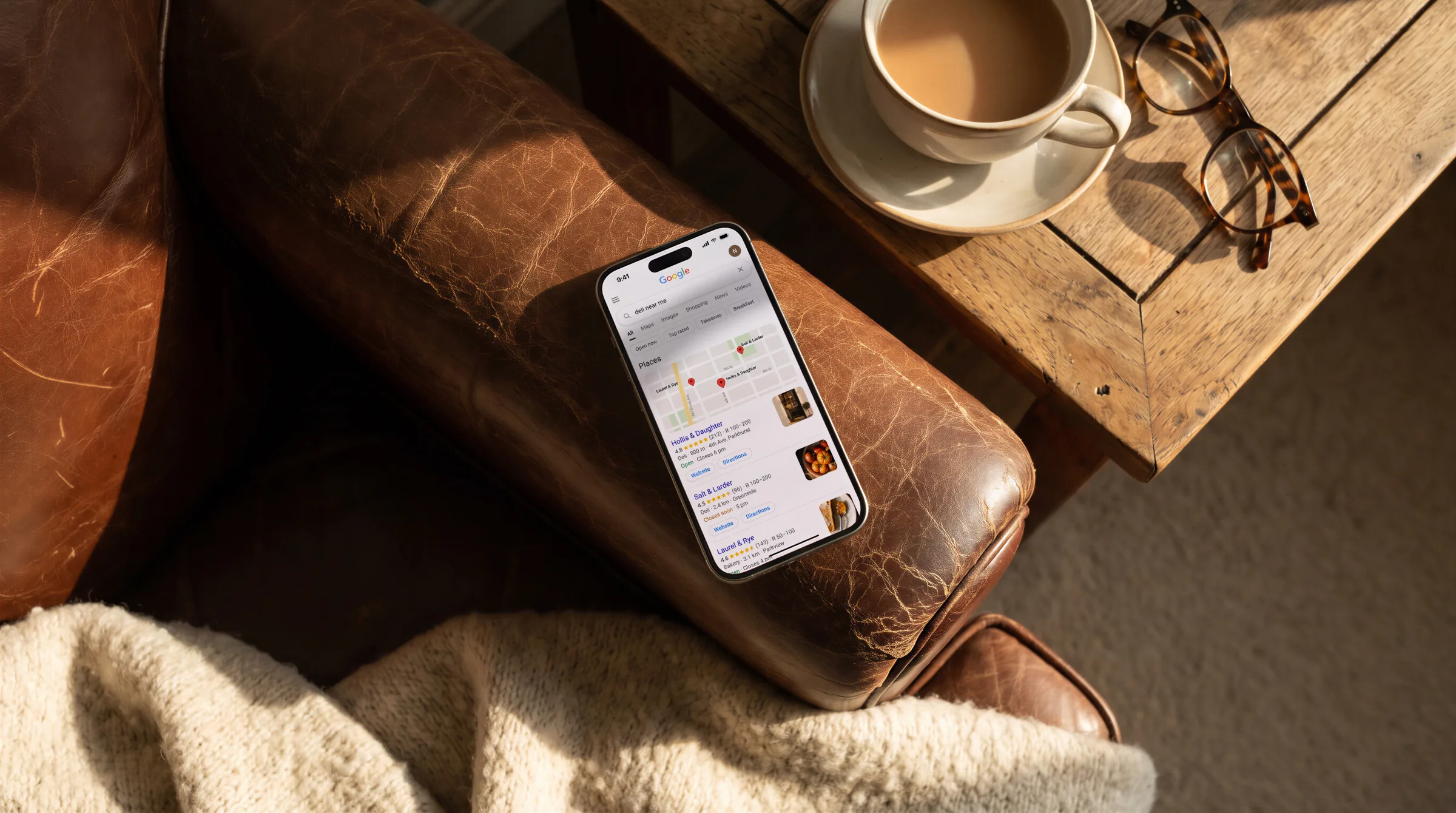 Google’s results for “deli near me”, a deli listed first, on a phone on a leather armchair’s arm beside tea and glasses.