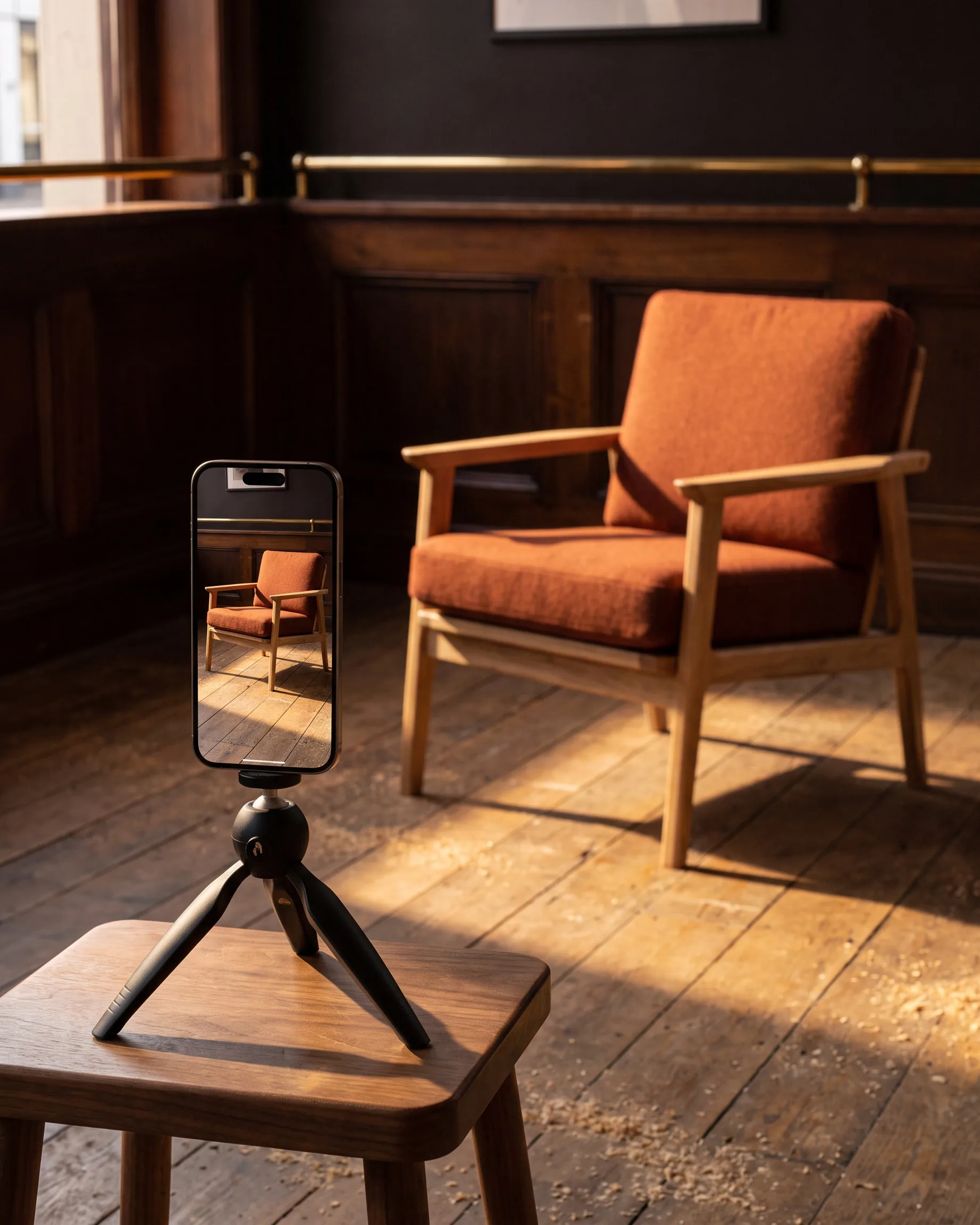 A phone on a tabletop tripod on a walnut stool filming a rust armchair in the sun, the chair live on its screen.