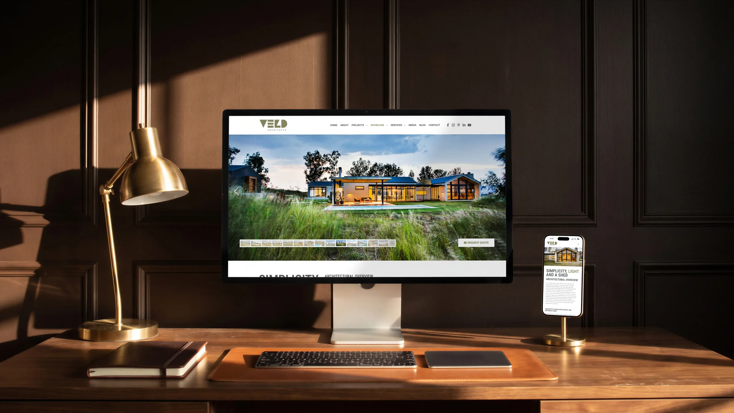 The same Veld Architects page on a desktop monitor and on a phone in a brass stand, on a walnut desk by a brass lamp.
