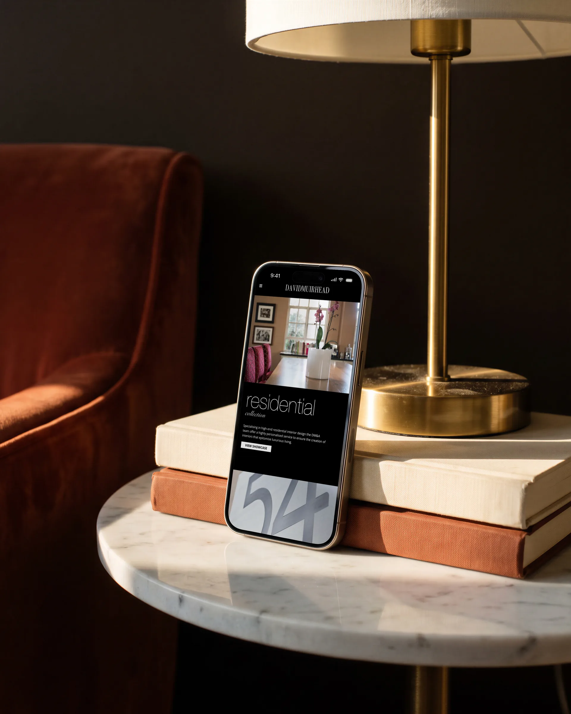 David Muirhead’s residential collection page on a phone leaning on books on a marble side table by a rust velvet armchair.