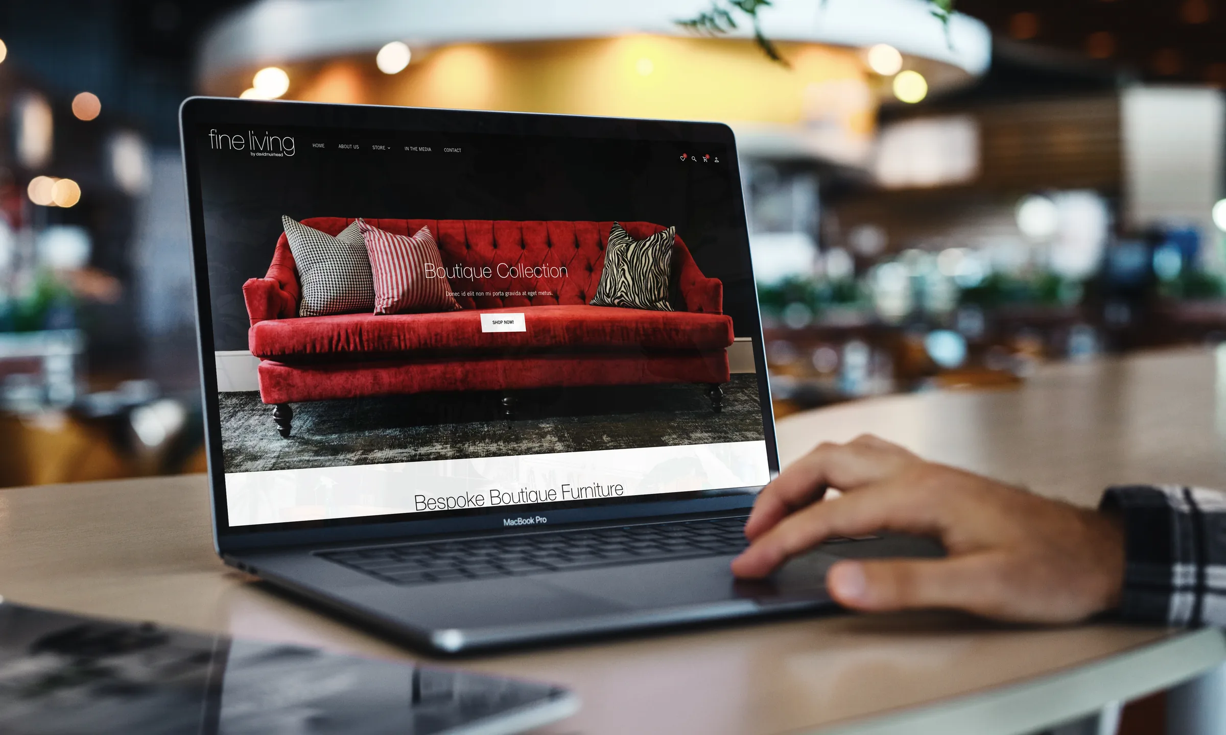 The home page on a laptop in a café: the red sofa under Boutique Collection, a hand on the trackpad.
