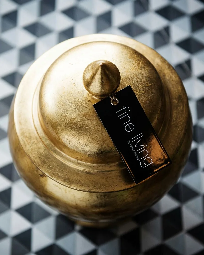 A black Fine Living swing tag hanging from a gold-leaf urn, over geometric tiles.