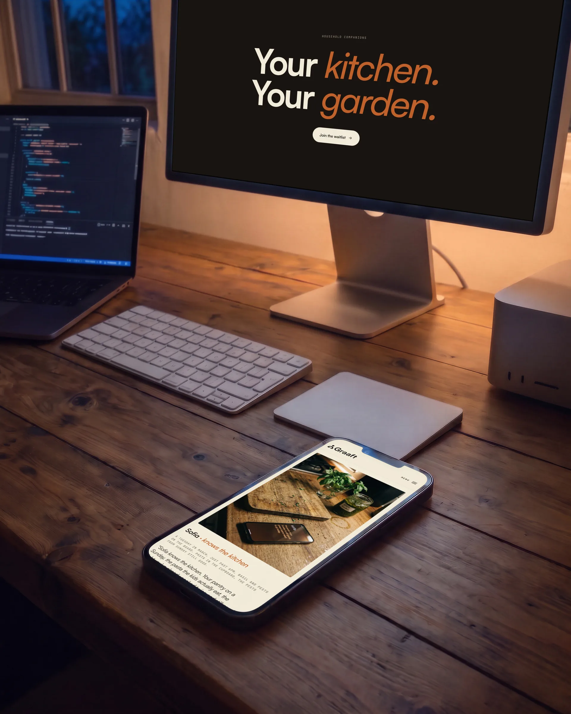 A desk at night from above: Your kitchen. Your garden. on the display, Sofia on a phone face up, a laptop showing code.
