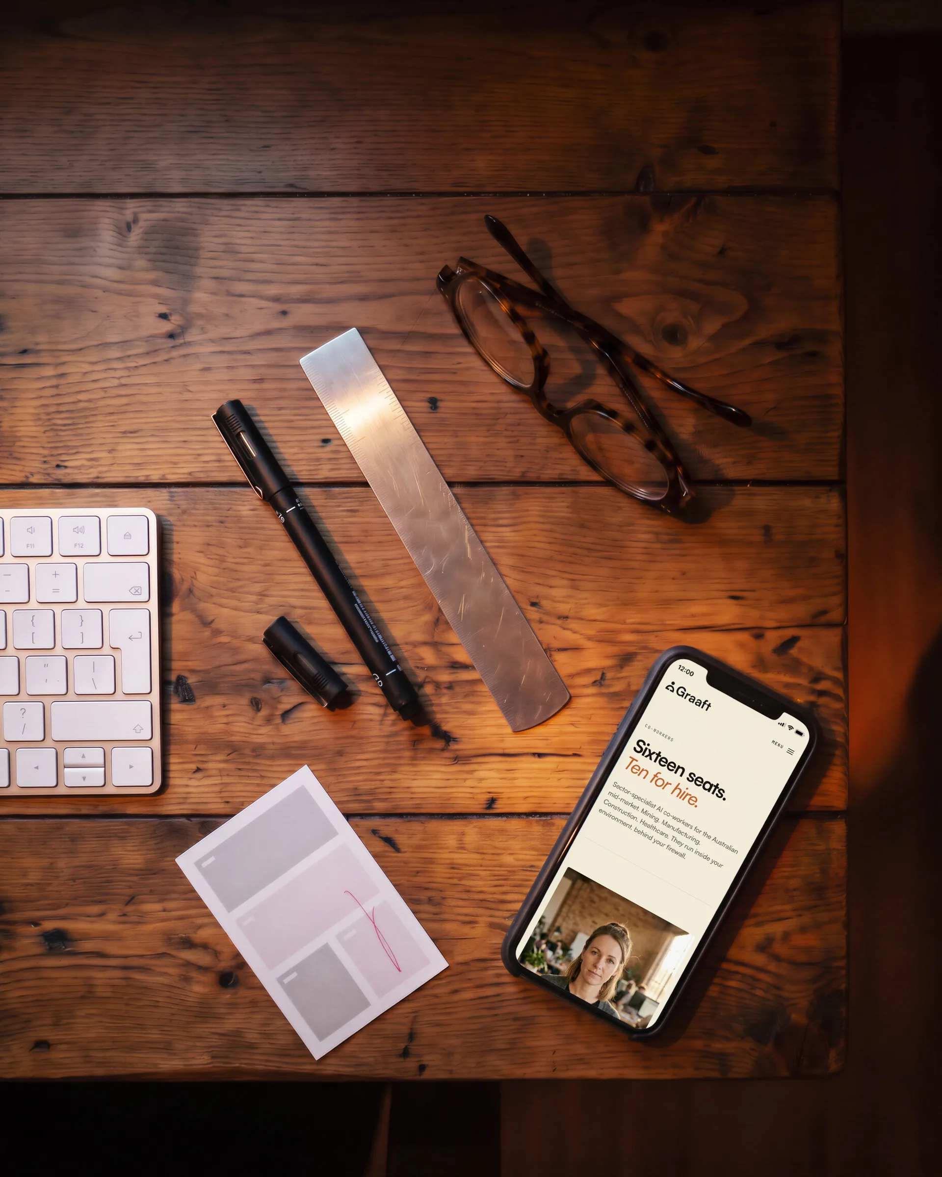 A timber desk from above: the co-workers page on a phone, Sixteen seats. Ten for hire., beside pens, a steel rule, glasses and a printed layout.