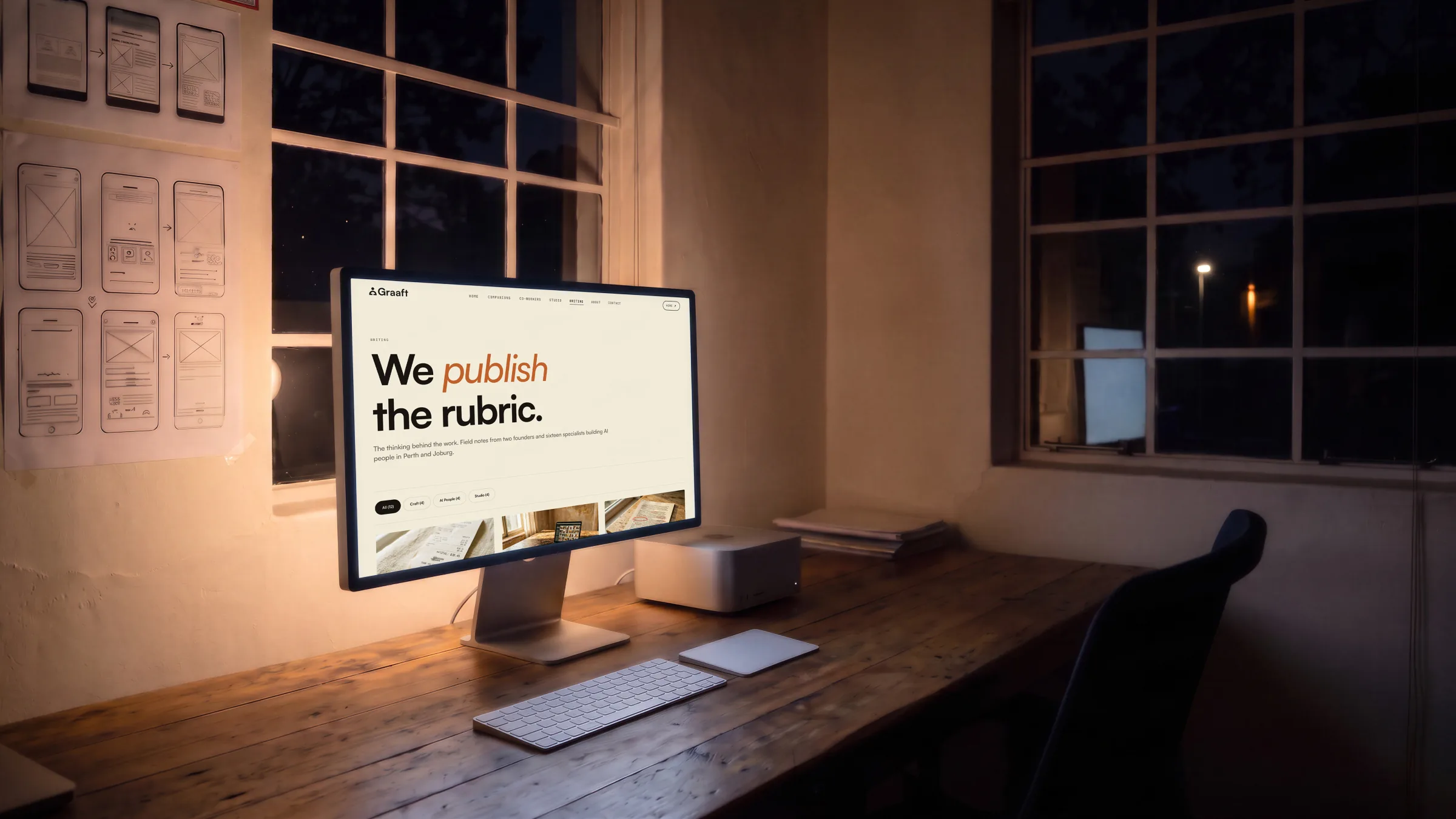 The studio at night: the Writing page, We publish the rubric., on a display on a long timber desk, wireframes pinned to the wall.